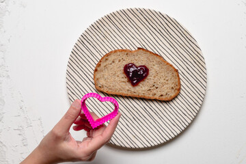 A hand holding a pink heart-shaped cookie cutter next to a slice of bread with heart-shaped jelly on a striped plate, over a white background.