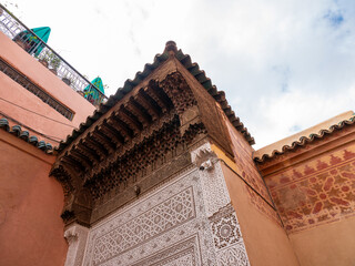 Detail of Ornate Moroccan Architecture in Marrakech, Showcasing Intricate Stucco Patterns and Carved Wooden Eaves Under a Cloudy Sky.