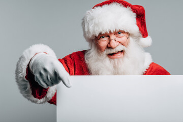 Naklejka premium Joyful Santa dressed in classic red pointing toward a blank signboard, expressing holiday cheer