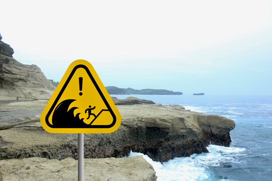 A yellow tsunami warning sign near the coast against a backdrop of blue sea, sky, and rocky cliffs or green forest. This coastal safety sign reminds people to remain cautious in tsunami-prone areas.