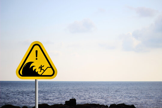 A yellow tsunami warning sign near the coast against a backdrop of blue sea, sky, and rocky cliffs or green forest. This coastal safety sign reminds people to remain cautious in tsunami-prone areas.