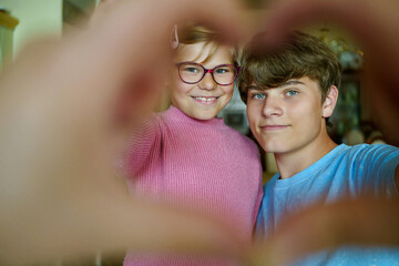 Happy teenage boy and little school girl making a heart shape with their fingers. Smiling siblings showing love, joy, and connection. Real life, real people.