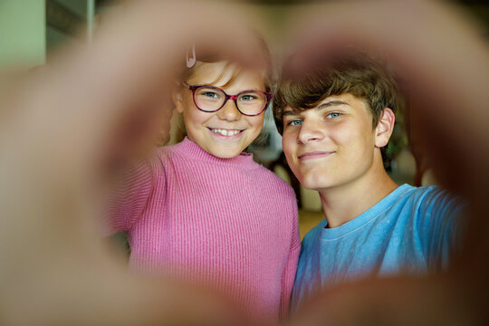 Happy teenage boy and little school girl making a heart shape with their fingers. Smiling siblings showing love, joy, and connection. Real life, real people.