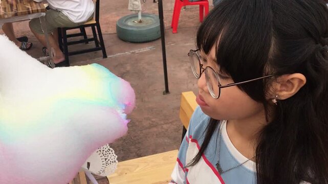Asian Teen Girl With Glasses Eating Cotton Candy At Park. A teen in specs bites soft pink fluff and smiles in a park vibe teen girl pink sweet
