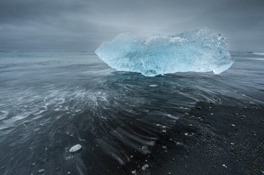 A shimmering blue iceberg is washed up by waves on a dark beach, Diamond Beach, J&ouml;kuls&aacute;rl&oacute;n glacier lagoon or J&ouml;kulsarlon, South Iceland, Iceland