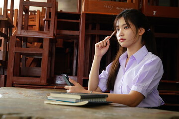 teenage Asian female student wearing a high school uniform sits reading a book and writing a homework report in a school classroom.