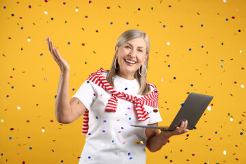 Excited senior woman with laptop and flying confetti against yellow background