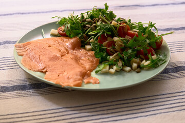 Top-down view of salmon slices and a fresh salad with arugula, cherry tomatoes, cheese, and pumpkin seeds served separately on an olive-colored plate. Healthy and cozy.