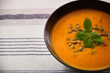 Close-up of creamy pumpkin soup in a black bowl on a rustic linen tablecloth, garnished with pumpkin seeds and basil. Cozy autumn meal, perfect for seasonal cooking.