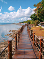 Naklejka premium A serene wooden walkway stretches along the shoreline, bordered by lush greenery and vibrant clouds, leading to a peaceful coastal view.