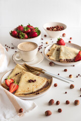 Crepes with chocolate hazelnut spread, strawberries and hazelnuts on a white wooden table.