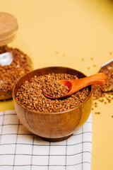 Buckwheat groats in a wooden bowl on a yellow background with a spoon and jar nearby