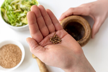 Whole coriander seeds on hand palm. Wooden mortar, avocado and guacamole ingredients background. Home cooking fresh spices. Aromatic natural authentic traditional Mexican homemade culinary atmosphere.