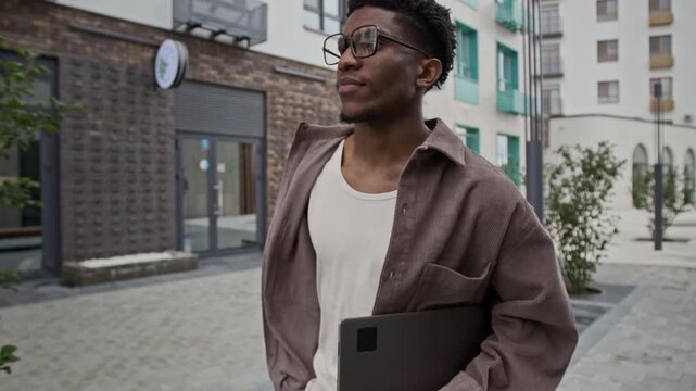 Close up of African American digital nomad carrying laptop while walking in downtown with modern buildings