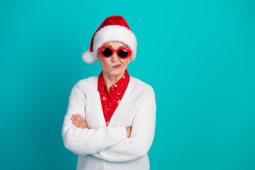 Confident elderly woman wearing a Santa hat red sunglass and white cardigan poses with arms crossed...