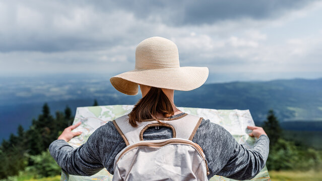 Woman hiker in sun hat reading a map on mountain ridge, rear view navigation and route planning with panoramic landscape under dramatic cloudy sky, outdoor travel and adventure concept - Powered by Adobe