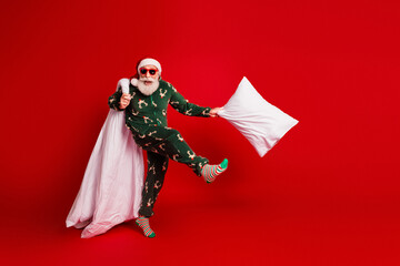 Cheerful santa in green pajama kicks a pillow on a bold red studio background creating a playful festive christmas mood