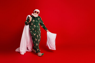 Naklejka na ściany i meble Santa in green christmas pajama with white pillow strikes a playful pose on a red background for cozy festive family and holiday marketing