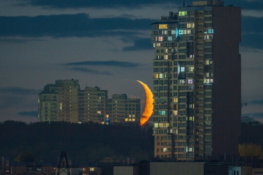 Crescent moon setting behind tall city buildings at night. Urban skyline with glowing lunar edge, illuminated windows and dark sky create a dramatic nightscape.