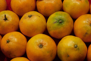 Mandarins with visible natural textures and color variations arranged closely on a red surface. The image evokes themes of local produce, healthy eating, affordability, and family economy.