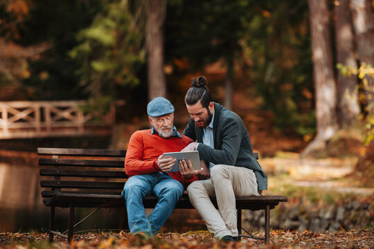 Senior father and his son sitting on bench by lake in nature, looking at tablet.