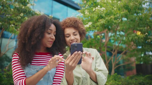 Two African American females sitting outdoors near trees holding smartphone together. Smiling while shopping online and discussing purchases during coffee break in modern business area.