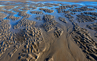 Tidal creeks at low tide on the sandy beach of Norderney island (Germany) flowing towards the North Sea and currents forming meandering temporary river beds. Blue sky mirroring in water surface.