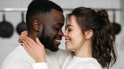 Happy interracial couple embracing intimately in kitchen, touching noses and smiling. Young man and woman in white sweaters sharing romantic moment, love and affection. Valentine's day relationship.