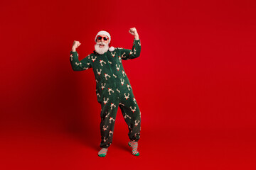 Celebratory Santa in green pajama suit dances with joy against red background for Christmas...