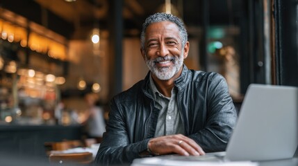 Obraz premium A joyful man with gray hair smiles while using his laptop in a warm cafe atmosphere.