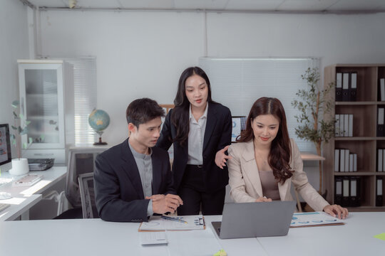 Three young Asian business professionals collaborate around a laptop and documents in a modern office, discussing strategy, analyzing reports and planning a project together