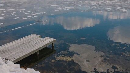 A metal dock is on a snowy shore with an icy blue sky reflected in the mirror-like surface of a partially frozen lake 