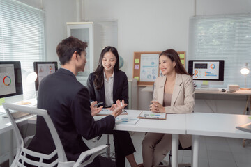 Three smiling Asian business professionals collaborate at a desk, reviewing charts and reports on monitors and paper, conveying teamwork, strategy, and successful communication in a modern office