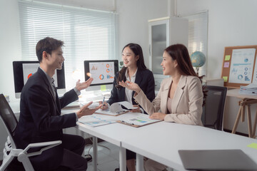 Business professionals collaborating at an office meeting, reviewing charts and graphs on computer monitors and documents, actively engaging in discussion about project development and strategy