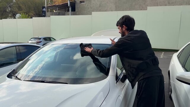 A young man cleans the exterior of a sedan car in a sunny outdoor parking lot. He is focusing on the car's surface using a soft, black microfiber cloth.