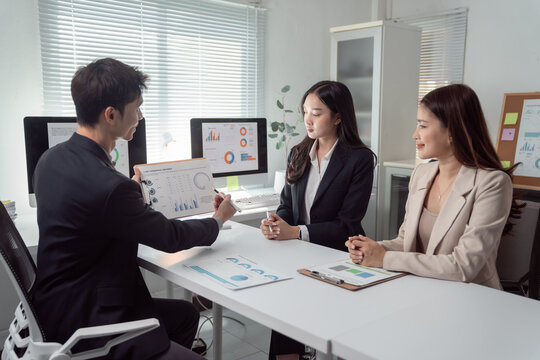 Business team members collaborating and discussing financial data charts and graphs on a clipboard and computer screens during an important meeting in a corporate office setting