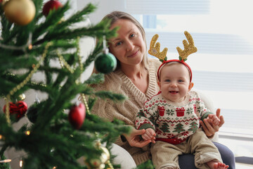 Mother with her baby wearing reindeer horns by the Christmas tree.
