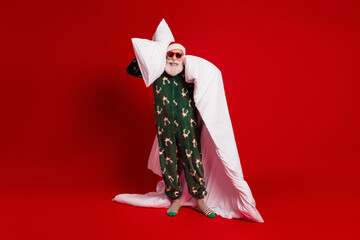 Naklejka na ściany i meble Cheerful Santa in green pajama with reindeer print holds white pillows and a long blanket against a bold red backdrop creating a playful holiday mood