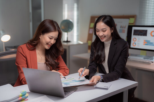 Businesswomen working together, smiling while analyzing financial reports and data on a laptop and monitor, demonstrating teamwork and professional collaboration in their office