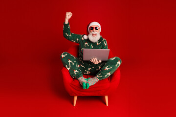 Naklejka na ściany i meble Santa in green Christmas pajama sits in red armchair with laptop joyfully celebrating online shopping and holiday fun