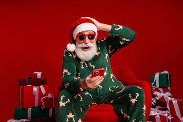 Naklejka na ściany i meble Happy Santa in festive pajama holds a smartphone with gifts and a warm red backdrop capturing Christmas cheer and holiday joy