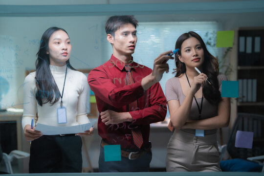 Asian business professionals collaborating, brainstorming and planning new ideas, writing notes and discussing strategies on a transparent glass board in a modern office environment