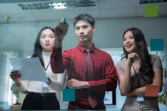 Asian business team collaborating around a glass whiteboard, brainstorming marketing strategy and planning projects in a modern office, focused, creative and confident - Powered by Adobe