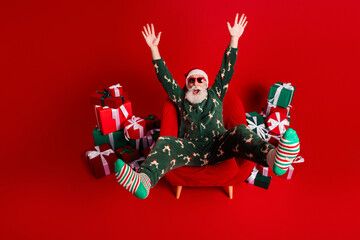 Naklejka na ściany i meble Cheerful Santa in festive pajama suit sits in red chair surrounded by gifts with striped socks celebrating christmas eve at home