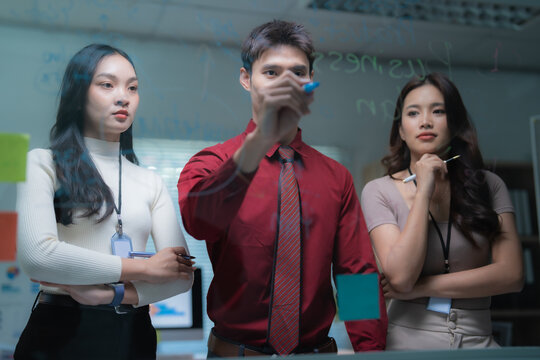 Young Asian business professionals actively collaborating, brainstorming ideas, and strategizing by writing on a clear glass board in a modern office meeting - Powered by Adobe