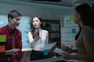 Business professionals collaborating and brainstorming ideas, writing on a transparent glass whiteboard during a late-night meeting in a modern office setup