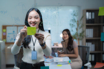 Asian businesswoman organizing ideas and brainstorming new project strategies using post-it notes on a transparent glass board in a dynamic modern office environment