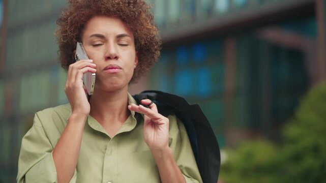 African American woman talking on smartphone outdoors near office building. Female exhaling after hearing disappointing news. Holding jacket on shoulder during conversation outside.