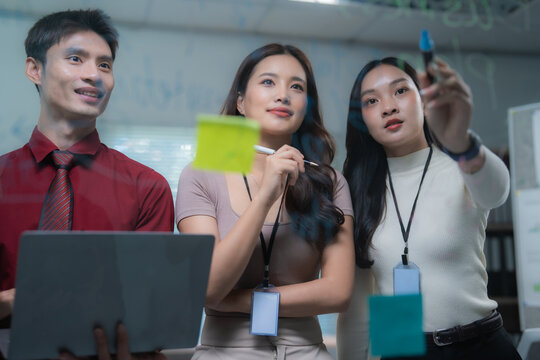 Asian business team collaborating and brainstorming new ideas, writing notes and discussing strategy on a clear glass board in a modern office meeting, demonstrating teamwork