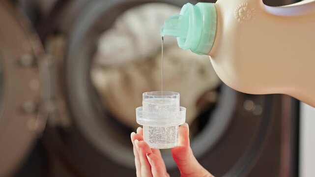 A woman pours liquid laundry detergent into a cap. Close-up of a woman's hands holding a measuring cap and pouring laundry gel, with a washing machine in the background. Laundry Detergent concept.